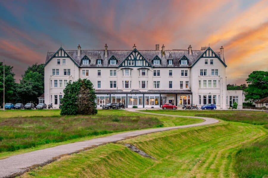 The Dornoch Hotel with a white facade and Tudor-style detailing, set against a colorful sunset sky, with a winding path leading up to the entrance and parked cars in front. This image highlights accessible travel in Scotland.