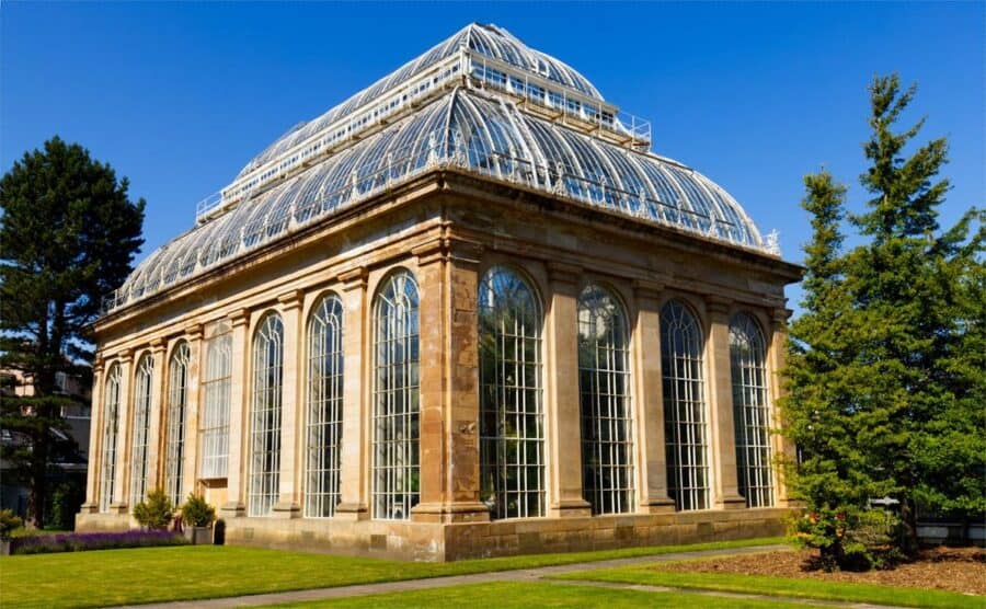 Glasshouse at the Royal Botanic Garden Edinburgh, featuring a grand Victorian-style structure with tall arched windows and intricate metalwork, set against a backdrop of lush greenery and a clear blue sky.