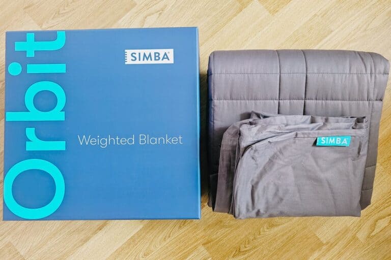 A blue box labeled "SIMBA Orbit Weighted Blanket" sits next to a folded dark gray SIMBA weighted blanket on a wooden floor. The blanket features a visible tag with the SIMBA logo.