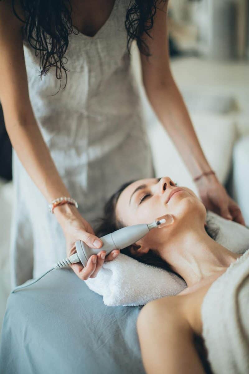 A woman receiving a laser resurfacing treatment in a spa-like setting. The clinician is using a handheld laser device on the woman's face, who is lying down with a towel under her head. The environment appears calm and serene, emphasising the relaxing aspect of the skincare treatment.