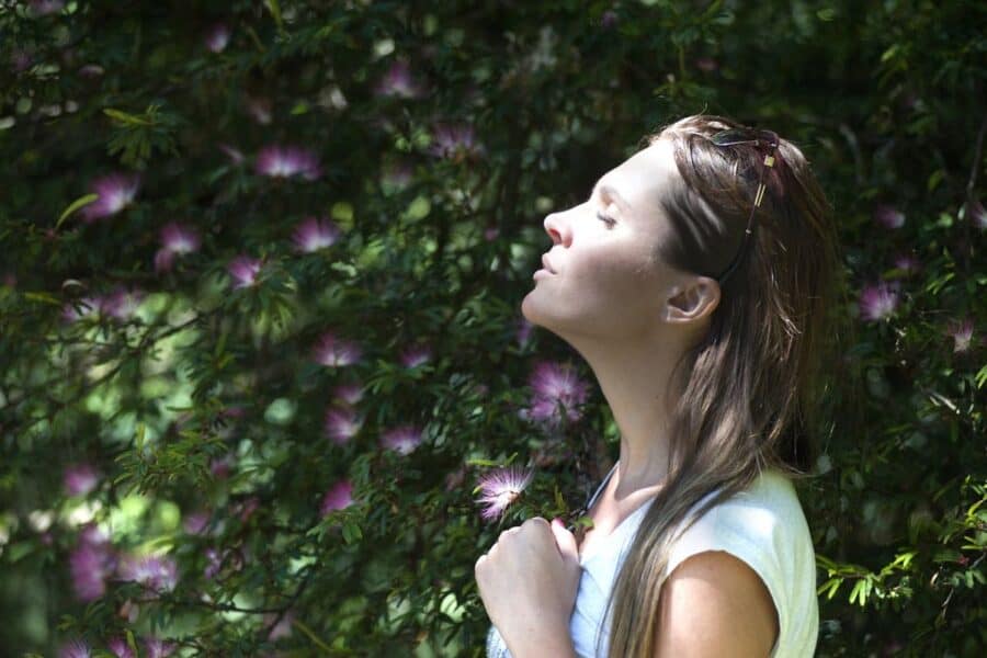A woman standing outdoors with her eyes closed and face turned towards the sun, surrounded by lush green foliage and blooming pink flowers. She appears to be enjoying the sunlight, with sunglasses resting on her head and a serene expression on her face.