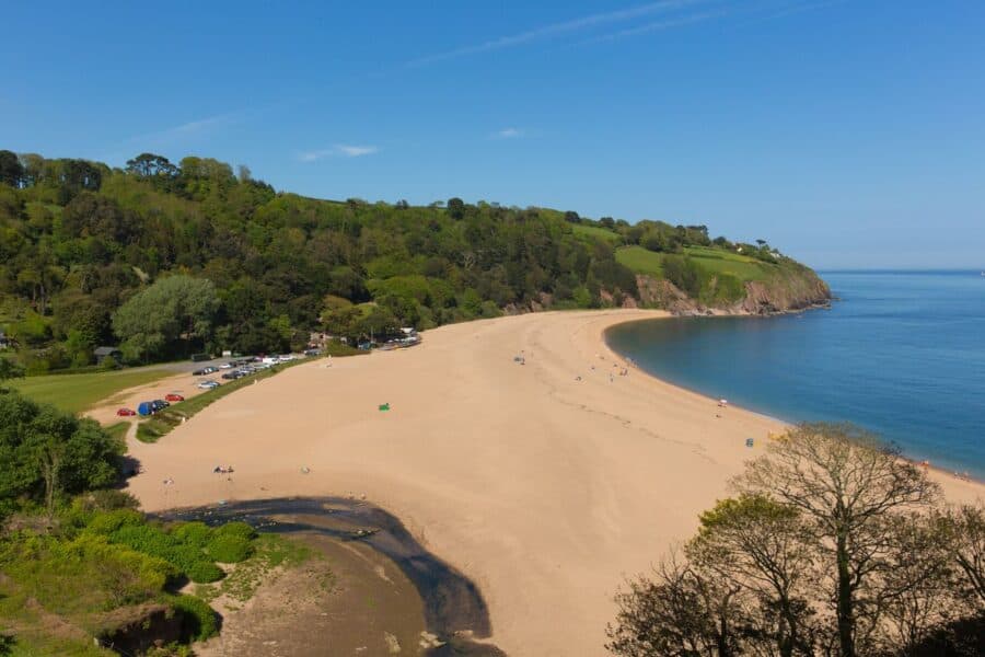 Aerial view of Blackpool Sands in Devon, a sandy beach nestled in a cove surrounded by lush green hills and a clear blue sea, with a few visitors scattered across the beach.