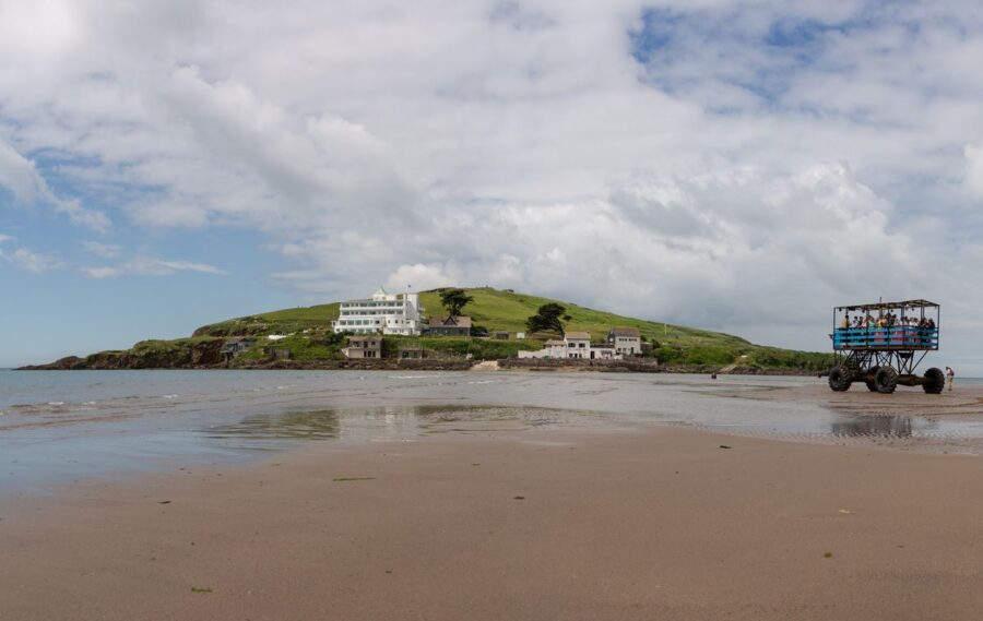 A view of Burgh Island in Devon with the Art Deco Burgh Island Hotel on a hill, and a large sea tractor carrying passengers across the beach.
