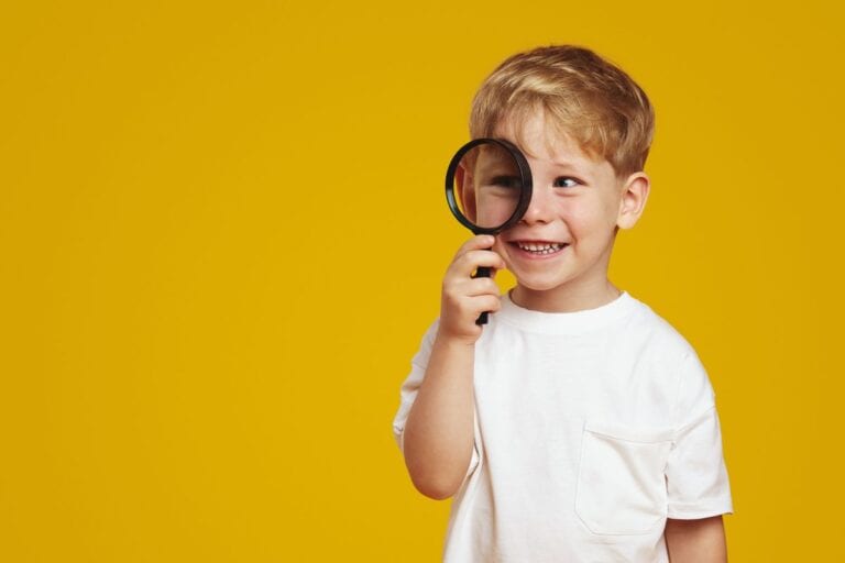 A smiling young boy in a white tshirt, against a mustard yellow background, holding a magnifying glass up to his eye. A great item to help encourage curiosity in children.