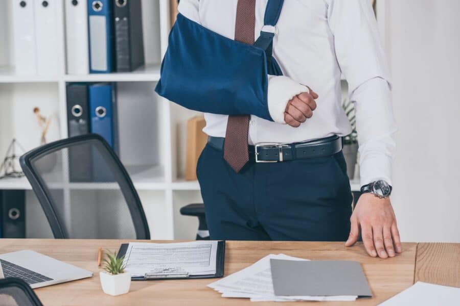 A man with an arm in a sling stands in an office, reviewing paperwork related to returning to work after a workplace injury.