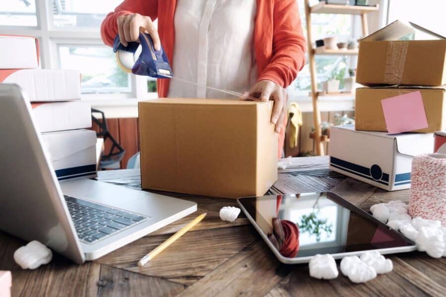 Person sealing a cardboard box with packing tape on a table surrounded by other packages, a laptop, tablet and packing materials, depicting a work environment related to e-commerce logistics and shipping.