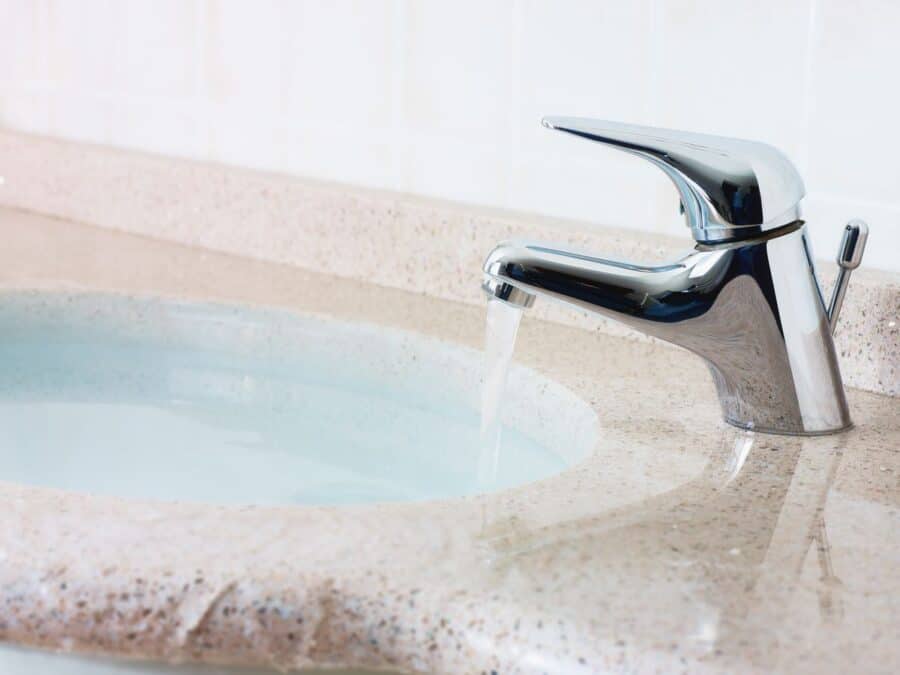 A chrome bathroom tap with water running into a clean, light-coloured sink on a speckled countertop.
