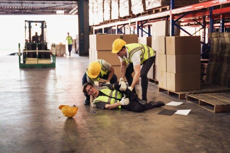 Two workers in safety vests and hard hats assist an injured colleague lying on the warehouse floor. A fallen hard hat and scattered papers are visible nearby, highlighting the aftermath of a workplace accident.
