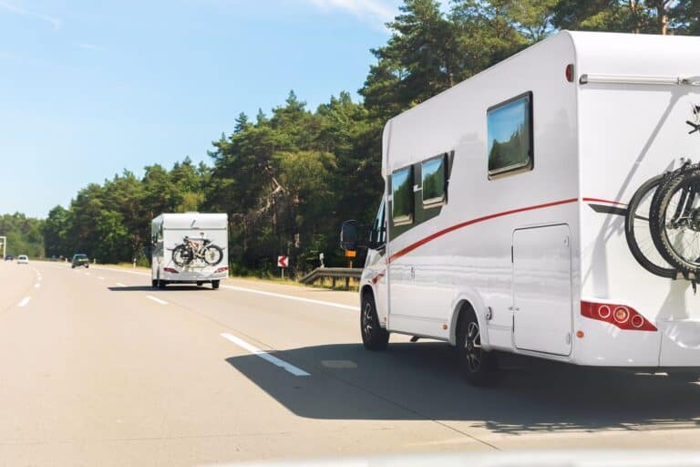 Two white motorhomes driving on a sunny highway, one carrying bicycles mounted on the back, surrounded by lush green forest scenery.