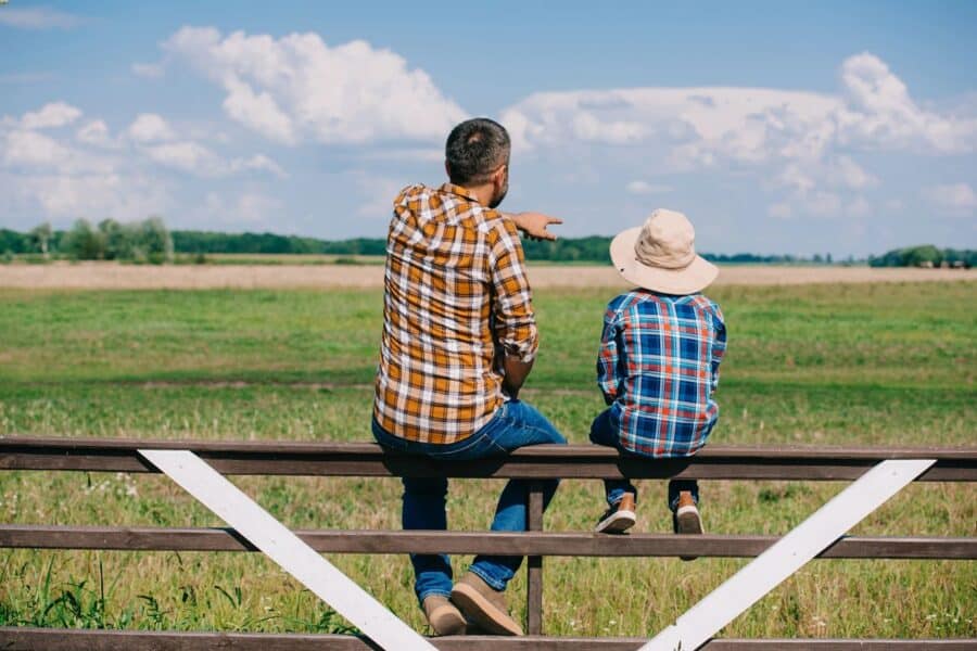A father and child sitting on a fence in a rural area, with the father pointing out towards the open field.
