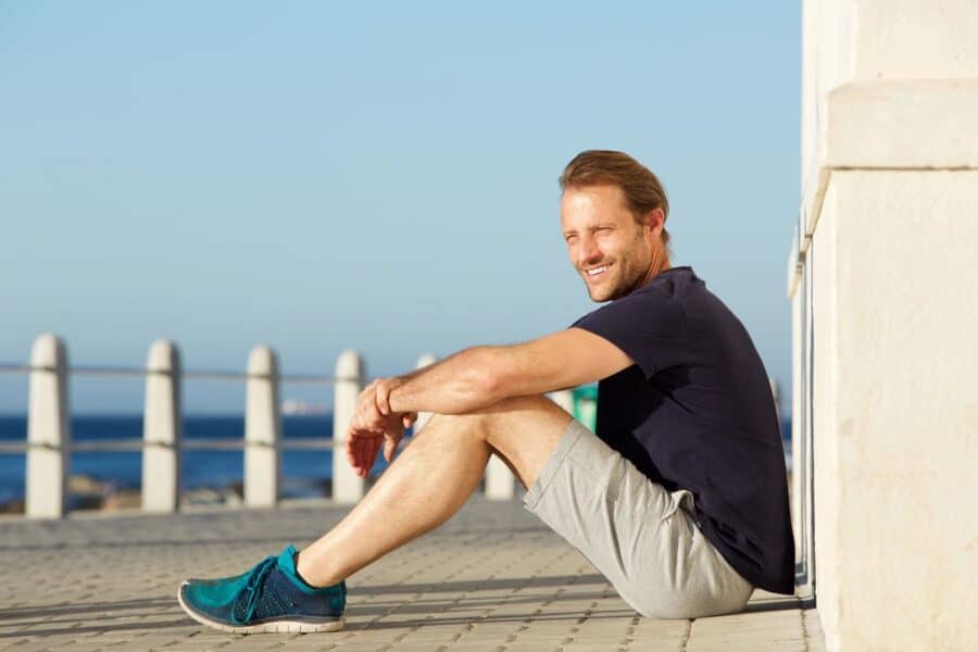 A man wearing light gray shorts and teal sneakers sits on a sunny seaside promenade, showcasing casual and functional menswear for winter sun destinations.
