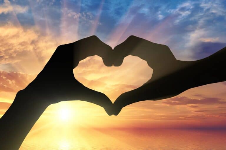 Silhouette of hands forming a heart shape against a vibrant sunrise over a calm ocean, symbolising health and wellness.