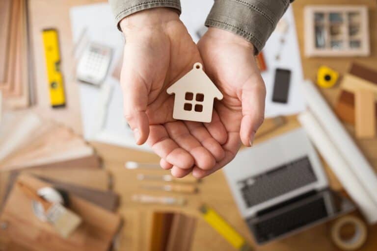 Hands holding a small wooden house model over a workspace filled with renovation tools and materials, highlighting the importance of house insurance during renovations.