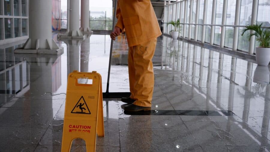 A maintenance worker in an orange uniform mopping a wet floor inside a modern, glass-walled corridor. In front of them, a yellow caution sign warns of the wet floor.
