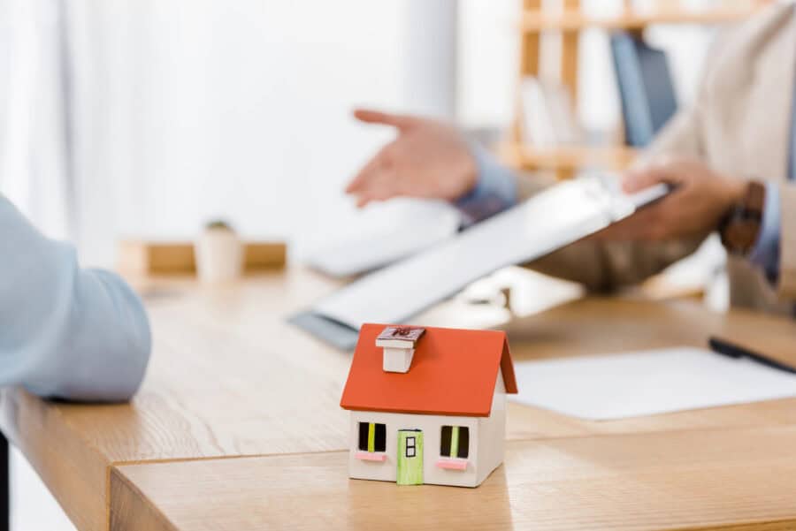 A miniature house model with a red roof on a wooden desk, while two people discuss documents in the background, illustrating the process of reassessing house insurance during renovations.