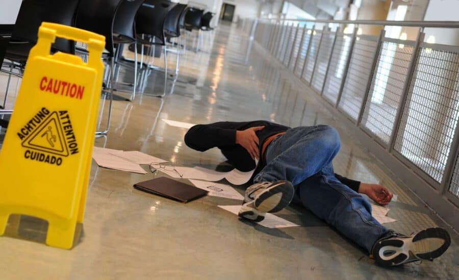 A person lying on the floor after a fall in a public corridor next to a yellow "Caution" sign.