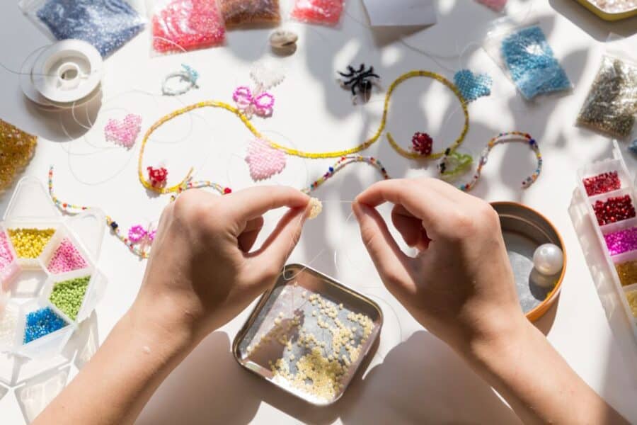 Hands threading small yellow beads with various colorful beads, tools, and handmade jewelry pieces scattered on a white surface, showcasing new ways to be creative.