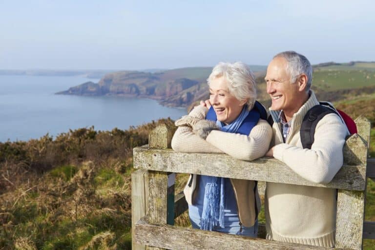 A smiling senior couple leaning on a wooden fence overlooking a scenic coastal landscape, representing the changing retirement landscape with active lifestyles and nature-focused leisure.