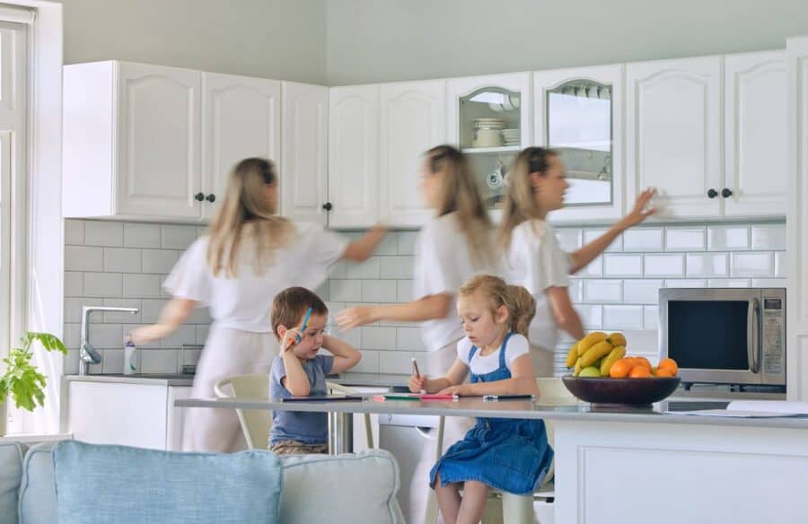A busy parent multitasking in the kitchen while two children sit at the counter doing homework, symbolising the daily challenges of parenting.