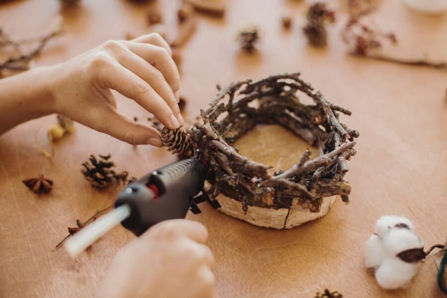 Hands using a hot glue gun to attach a pine cone to a small decorative nest made of twigs, showcasing new ways to be creative with everyday items.