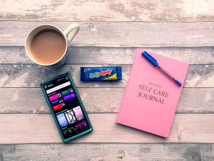 Flat lay of a self-care kit including a pink self-care journal, pen, smartphone, cup of tea and a chocolate bar.