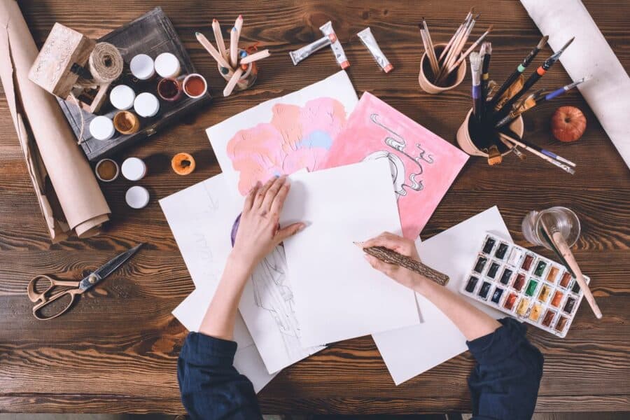 Artist’s hands working on a drawing at a wooden table surrounded by art supplies, sketches, paint jars, and brushes, showcasing new ways to be creative.