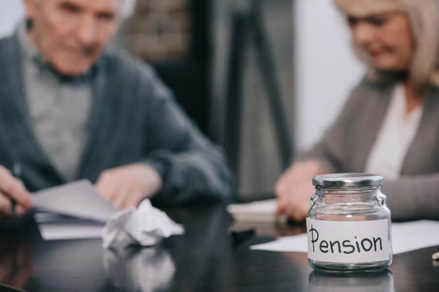 A glass jar labelled "Pension" sits on a desk, with two older adults blurred in the background, discussing paperwork, highlighting the financial considerations of the changing retirement landscape.
