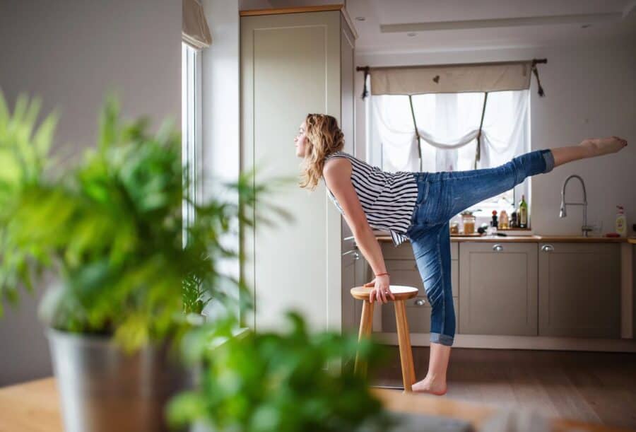 A woman practicing a balancing yoga pose in her kitchen, using a stool for support.