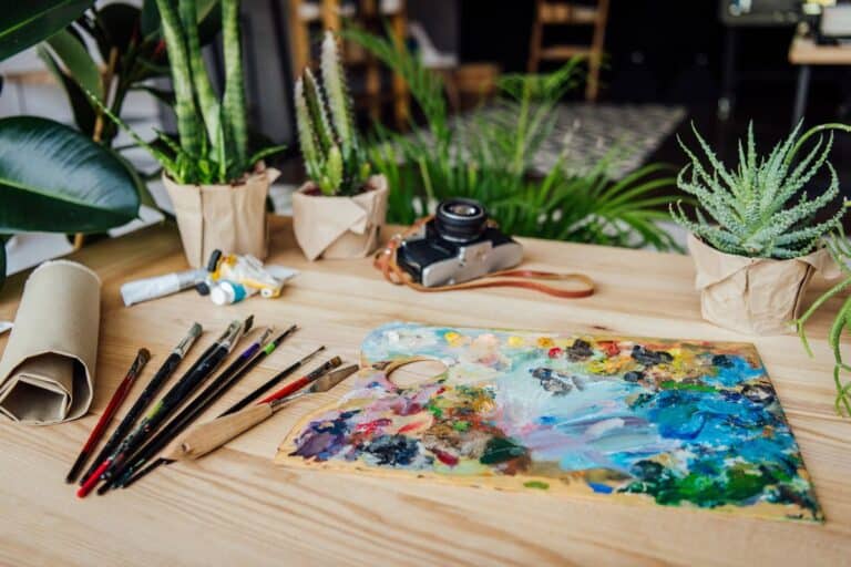 A creative workspace featuring an artist's paint palette, paintbrushes, and small potted plants on a wooden table, with a vintage camera in the background.