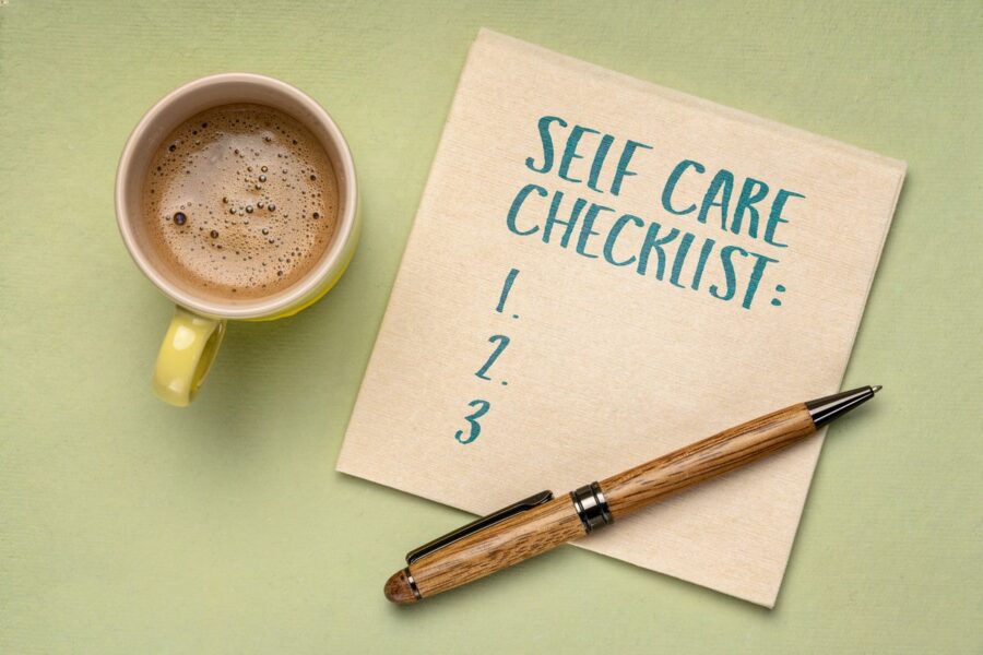 Flat lay image of a self-care checklist on a notepad next to a cup of coffee and a wooden pen, encouraging busy parents to share their self-care tips.