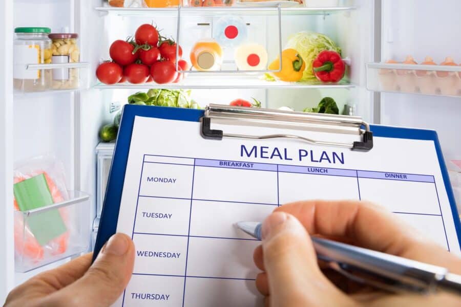 Close-up of a meal planning chart held in front of a fridge stocked with fresh ingredients as part of a Sunday evening routine.