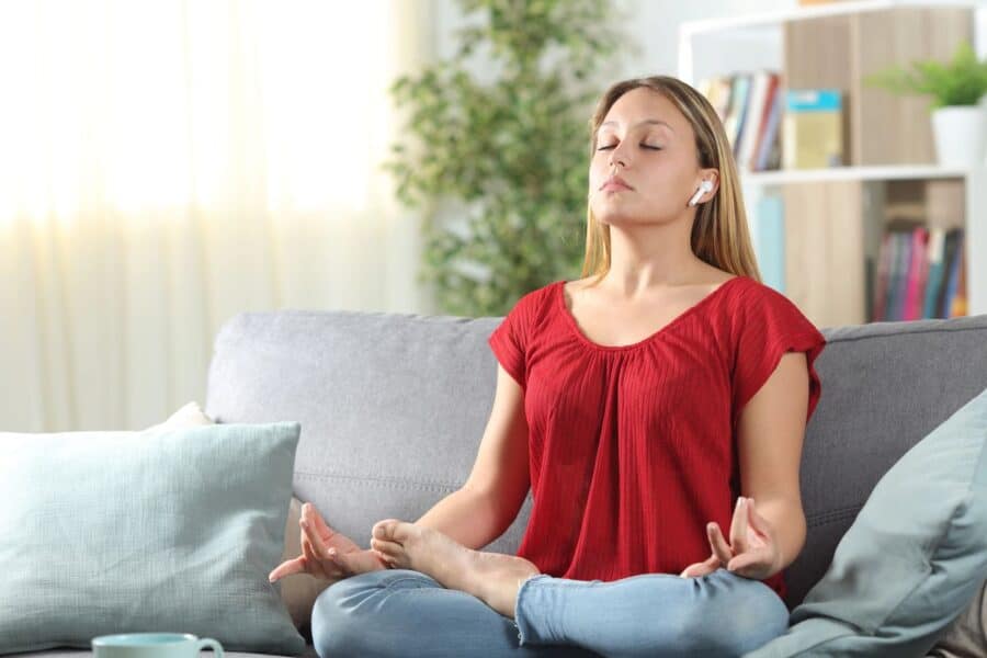 A woman practicing mindfulness on a sofa, sitting cross-legged with eyes closed, wearing a red top and earphones.