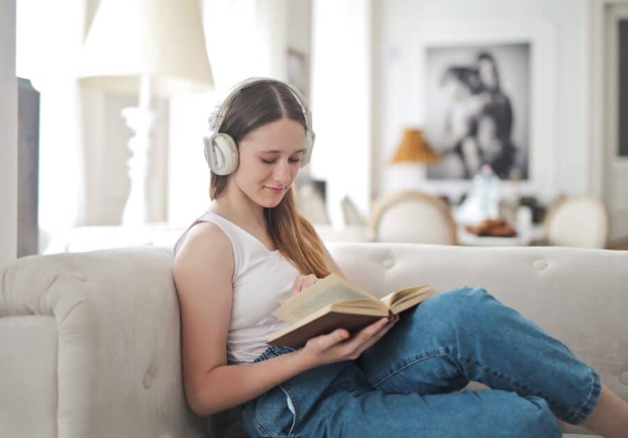 A young woman wearing headphones, relaxing on a couch while reading a book, creating a calm and peaceful atmosphere for a Sunday evening routine.