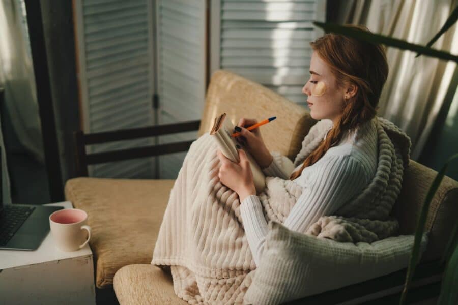 Young woman sitting in a cosy corner with a blanket, writing in a journal as part of a Sunday evening routine, with a cup of tea.