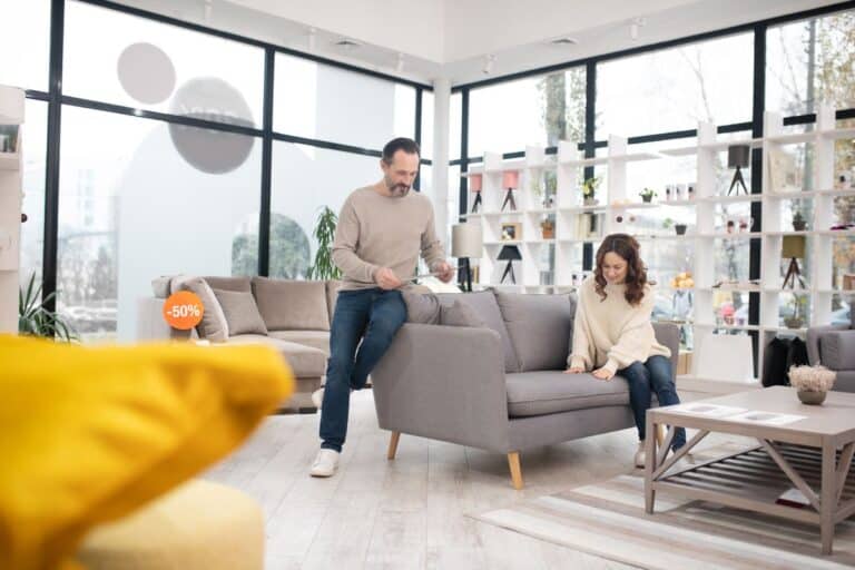 A woman sat on a sofa in a furniture store while a man sits on the arm of the sofa looking at the price.