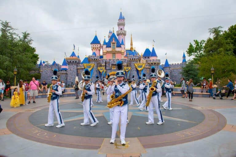 Disneyland marching band performing in front of Sleeping Beauty Castle at Disneyland Anaheim, with visitors watching in the background.