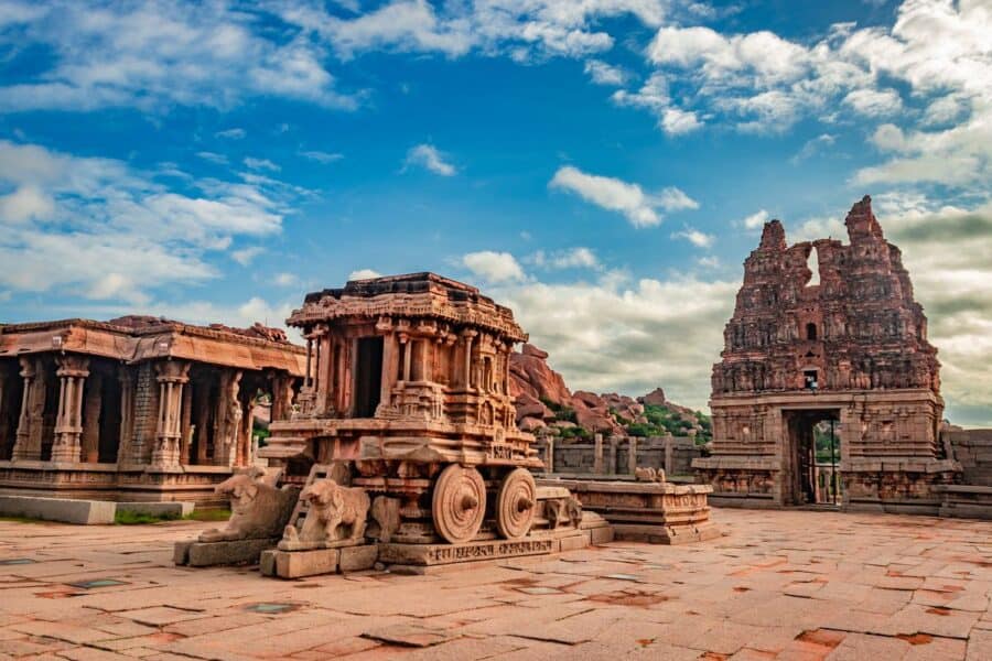 The iconic stone chariot and temple ruins at the Vittala Temple complex in Hampi, India, under a partly cloudy sky.
