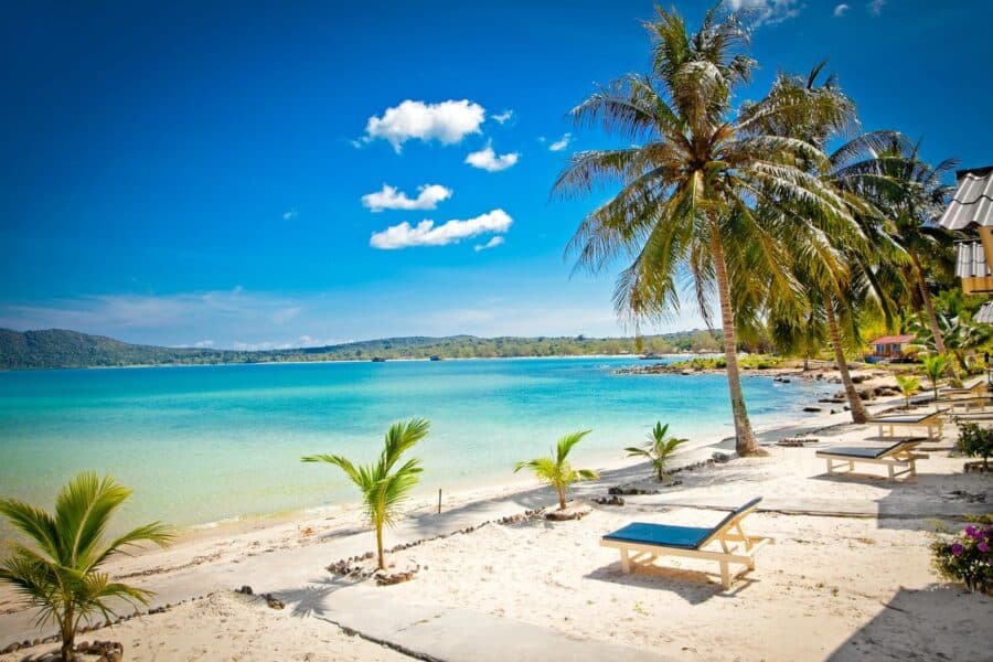 A tropical beach on Koh Rong Samloem, Cambodia, with clear turquoise waters, white sand, palm trees, and sun loungers under a bright blue sky.