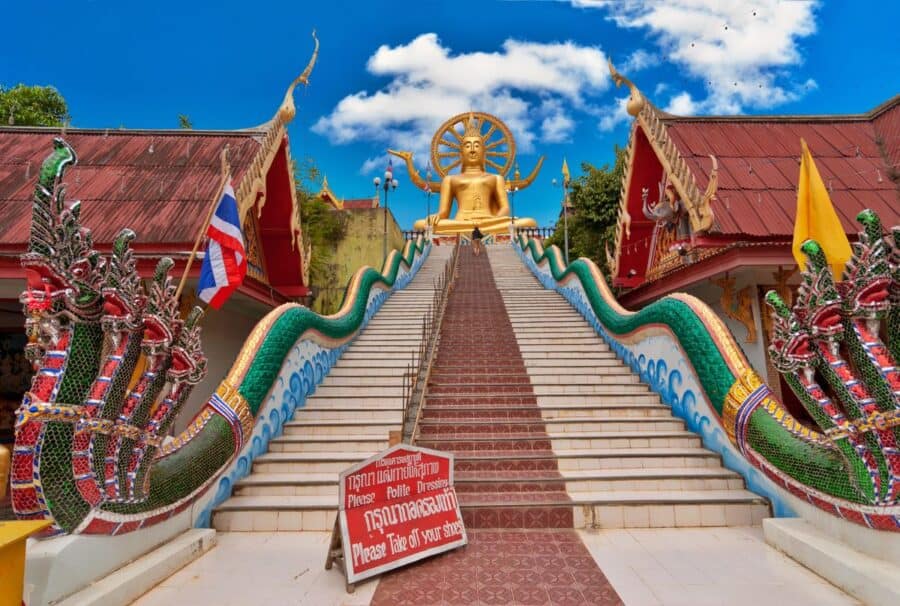 The Big Buddha statue on Koh Samui, Thailand, with a grand staircase flanked by colorful serpent sculptures under a bright blue sky.