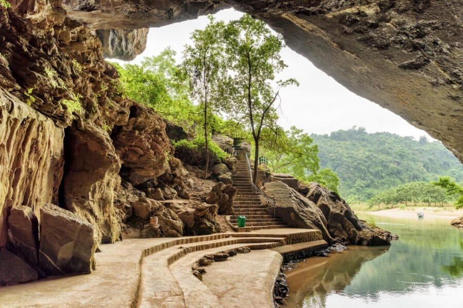 The entrance to a cave in Phong Nha-Ke Bang National Park, Vietnam, one of the many unhique destinations in Asia, with stone steps leading through lush greenery beside a calm river.