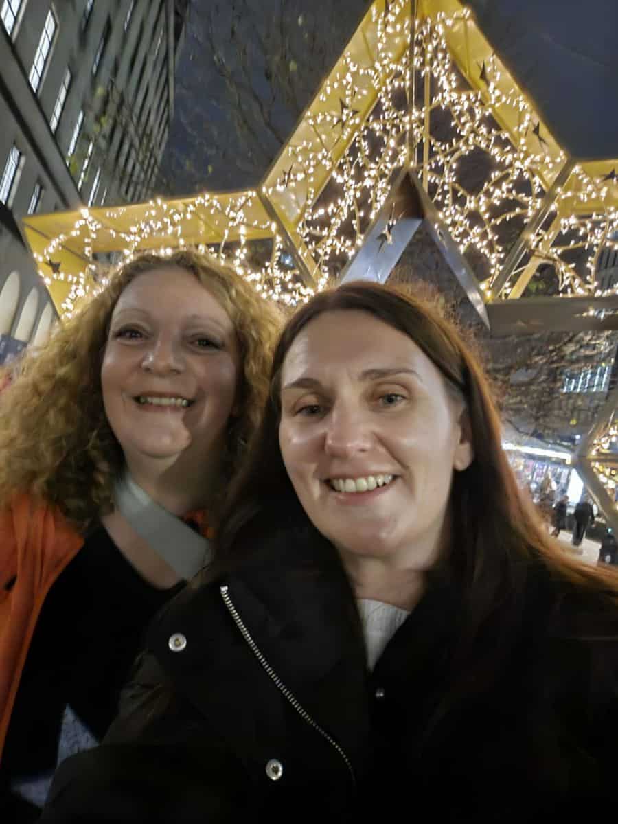 Two smiling women taking a selfie in front of illuminated star decorations at night in Manchester, representing a strong accountability partnership and support system for achieving goals.
