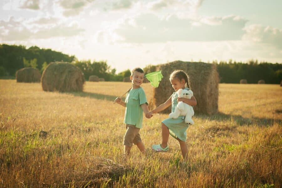 Two young children playing in a sunlit field, holding hands, with one carrying a stuffed toy and the other holding a butterfly net, promoting outdoor sustainable living