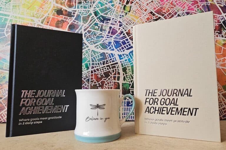 Two hardcover journals titled The Journal for Goal Achievement, one Graphite and one Sand coloured, are displayed against a colourful abstract map background. A white ceramic mug with a dragonfly illustration and the words "Believe in you" sits between them.