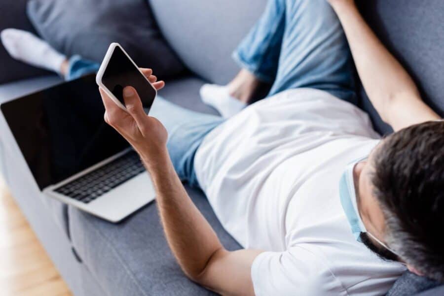 Man lying on a sofa browsing his phone with a laptop nearby.