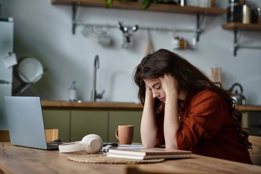 Tired young woman sitting at a kitchen table with her head in her hands, looking stressed next to an open laptop.