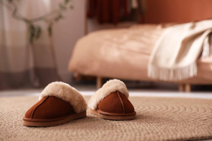 Shearling slippers on a woven rug beside a bed with a throw — bedroom reset for autumn, cosy textures and warm neutrals.