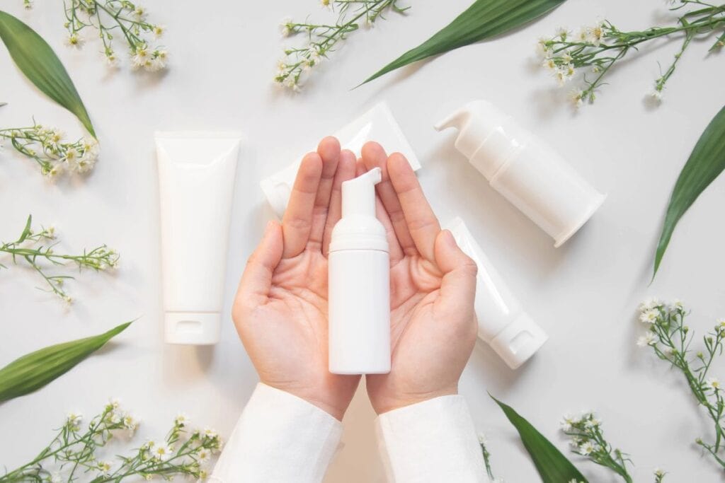 Hands holding a pump bottle with simple skincare tubes and leaves — essentials for how to build a skincare routine.