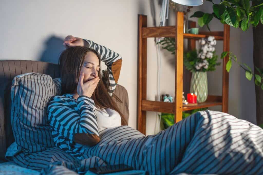 Woman yawning in striped pyjamas, winding down in bed with soft lamp and candle—calm bedtime routine for a better night’s sleep.