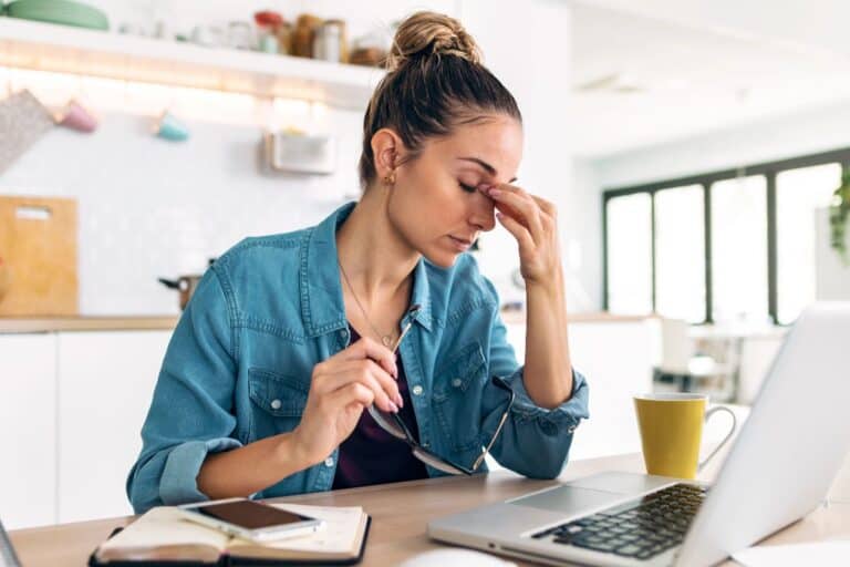 Stressed woman at laptop rubbing eyes in a kitchen, illustrating how to stop feeling overwhelmed with a quick reset.