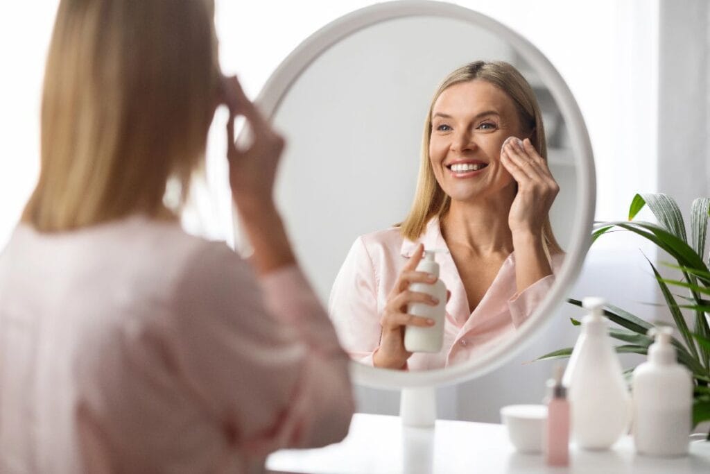 Smiling woman applying toner with a cotton pad in front of a mirror, refreshing your skincare routine with a simple daily step.